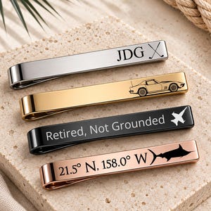 May include: Four rectangular metal tie clips in silver, gold, black, and rose gold tones. The silver clip features the text JDG and a golf club icon. The gold clip shows a car engraving. The black clip reads Retired, Not Grounded with a plane icon. The rose gold clip displays coordinates 21.5 N, 158.0 W and a fish icon.