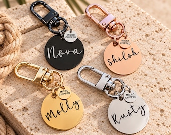 May include: Four circular metal pet ID tags with lobster claw clasps. Each tag features a name in script font and a smaller circular charm engraved with the text MICRO CHIPPED. The tags are finished in black, rose gold, gold, and silver tones.