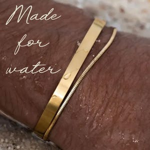 Pu&ograve; includere: Due braccialetti d'oro al polso di una persona. I braccialetti sono fatti di una catena sottile e di una fascia piatta pi&ugrave; spessa. Il testo "Made for water" &egrave; visibile nell'immagine.