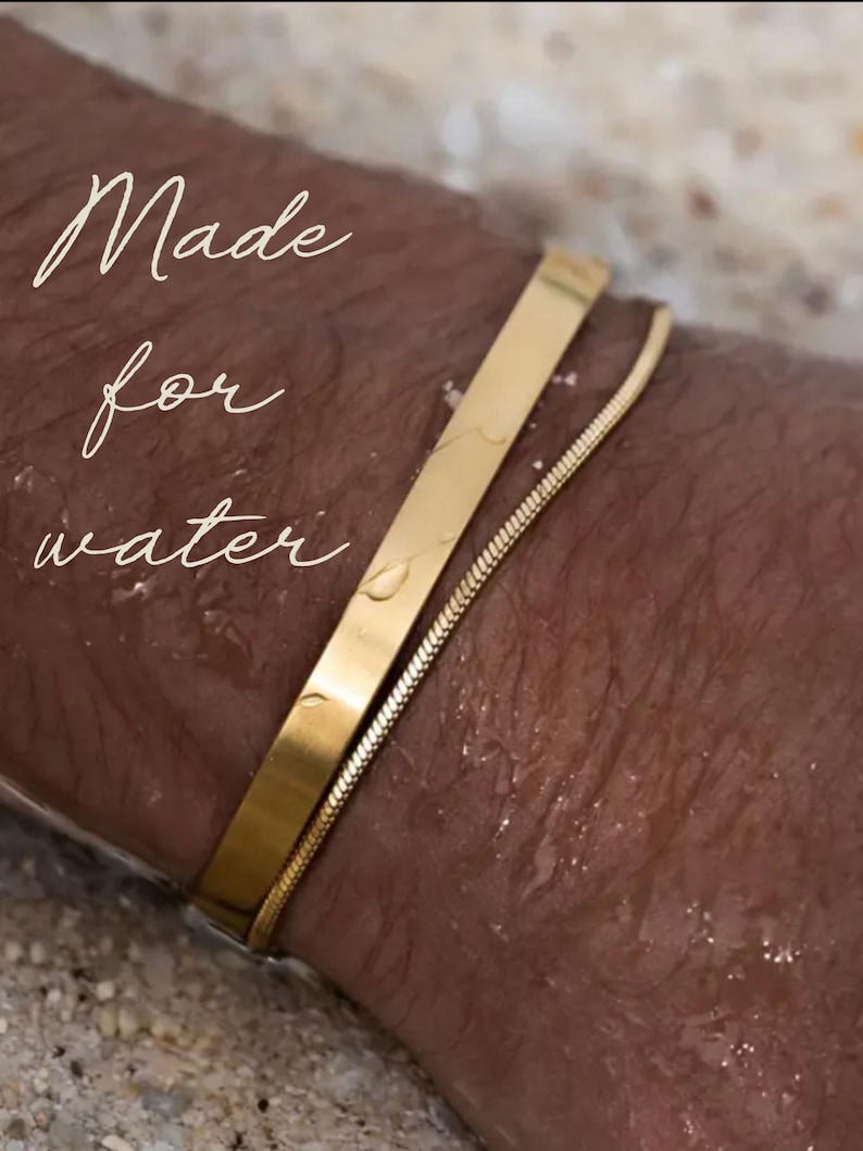 Pu&ograve; includere: Due braccialetti d'oro al polso di una persona. I braccialetti sono fatti di una catena sottile e di una fascia piatta pi&ugrave; spessa. Il testo "Made for water" &egrave; visibile nell'immagine.
