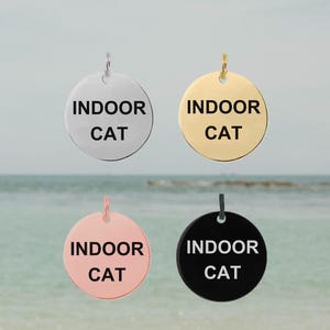 以下が含まれることがあります： 「INDOOR CAT」の文字が入った、丸いペットタグが4つ。シルバー、ゴールド、ローズゴールド、ブラックのタグがあります。各タグの上部には、首輪に取り付けるための小さなループが付いています。背景はビーチの風景です。