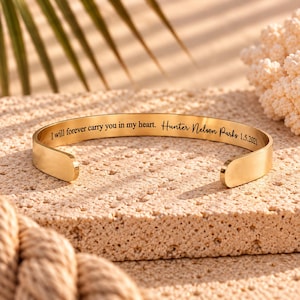 May include: A gold-toned metal cuff bracelet with a smooth, polished finish. The interior surface is engraved with the black text: I will forever carry you in my heart. Hunter Nelson Parks 1.5.2021. The bracelet features an open-ended design.