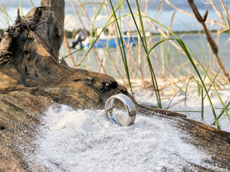 May include: A silver wedding band with an inscription on the inside of the band. The ring is lying on a white sandy surface with a piece of driftwood in the background.