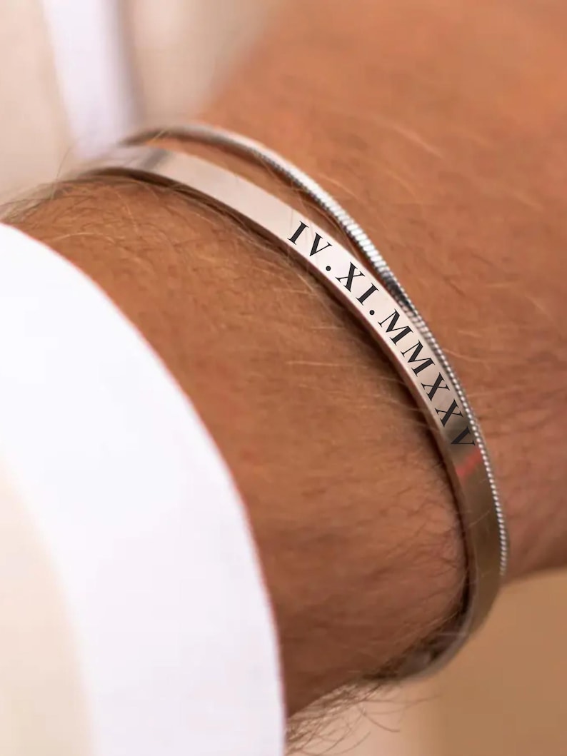 Pu&ograve; includere: Un braccialetto d'argento con una fascia sottile e una fascia pi&ugrave; spessa con numeri romani incisi. La fascia pi&ugrave; spessa recita "IV.XI.MMXV".