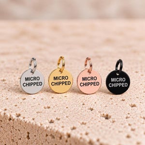 May include: Four circular metal pet tags with jump rings, each featuring the text MICRO CHIPPED in black capital letters. The tags are finished in silver, gold, rose gold, and black, arranged in a horizontal row on a textured surface.
