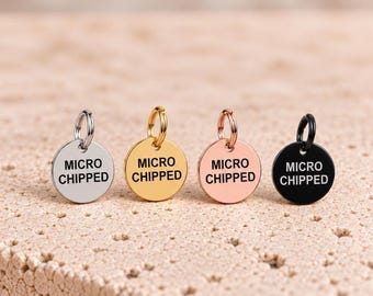 以下が含まれることがあります： 黒い大文字で「MICRO CHIPPED」と刻印された、4つの円形金属製ペットタグ。シルバー、ゴールド、ローズゴールド、ブラックの仕上げが施されており、それぞれに丸カンが付いています。タグはテクスチャのある表面に一列に並べられています。
