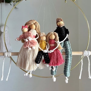 May include: A handcrafted family mobile featuring five figures made of wood and yarn. The figures are suspended from a gold-colored metal ring, with a wooden dowel at the bottom. The figures are dressed in various colors, including pink, yellow, black, and teal.