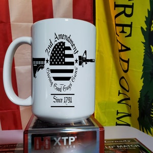 Pode incluir: Caneca de cerâmica branca com um design gráfico preto com uma bandeira americana, armas e o texto "2nd Amendment Giving Good Guys Since 1791". A caneca tem uma alça grande e está em uma caixa de balas.