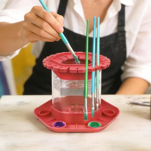 May include: A red paint brush cleaner and holder with a clear water reservoir. The holder has slots for brushes and small wells for paint. Several paint brushes are in the holder, and one is being dipped into the water.