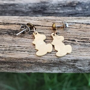 Puede incluir: Un par de pendientes de color dorado con una peque&ntilde;a silueta estilizada de un oso. Los pendientes tienen postes y respaldos plateados.