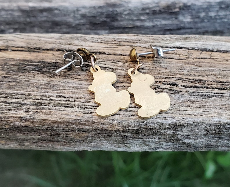 Puede incluir: Un par de pendientes de color dorado con una peque&ntilde;a silueta estilizada de un oso. Los pendientes tienen postes y respaldos plateados.