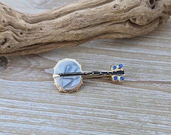 Puede incluir: Un broche de tono dorado con forma de banjo con un cuerpo de nácar blanco y detalles de pedrería azul.