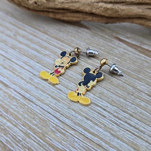 Puede incluir: Un par de pendientes de Mickey Mouse de tono dorado. Los pendientes presentan una imagen de dibujos animados de Mickey Mouse con pantalones cortos rojos y zapatos amarillos. Los pendientes est&aacute;n unidos a postes plateados.