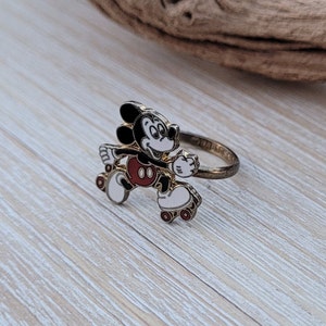 May include: A gold-tone ring featuring a cartoon image of Mickey Mouse roller skating. Mickey is wearing red shorts and white gloves. The ring is a fun and whimsical piece of jewelry.