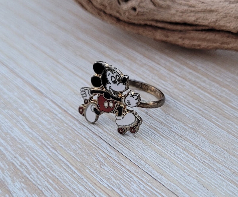 May include: A gold-tone ring featuring a cartoon image of Mickey Mouse roller skating. Mickey is wearing red shorts and white gloves. The ring is a fun and whimsical piece of jewelry.