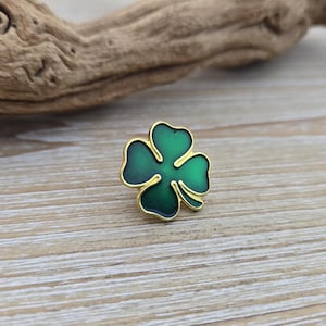 Puede incluir: Un broche de trébol de cuatro hojas verde con un borde dorado. El broche está sobre un fondo de vetas de madera clara. El trébol tiene un acabado brillante y es un símbolo de buena suerte.