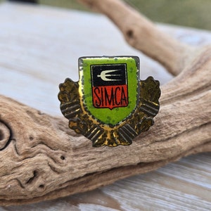 May include: A vintage metal pin with a green, red, and black shield featuring a stylized bird and the word "SIMCA". The shield is surrounded by gold-colored leaves.