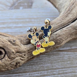 Puede incluir: Un par de pendientes de Mickey Mouse de tono dorado con detalles rojos y amarillos. Los pendientes est&aacute;n sobre una superficie de madera.