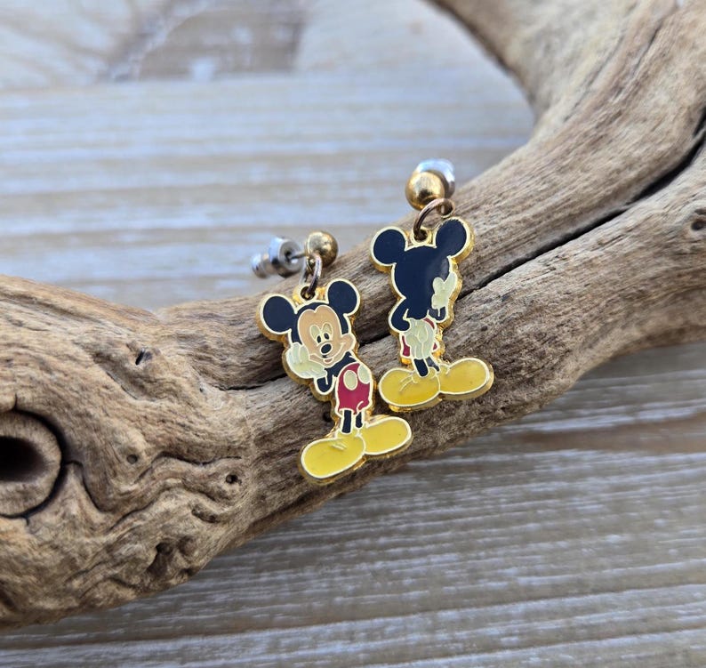 Puede incluir: Un par de pendientes de Mickey Mouse de tono dorado con detalles rojos y amarillos. Los pendientes est&aacute;n sobre una superficie de madera.