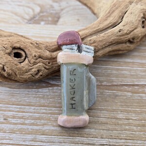 Puede incluir: Pequeña figura de cerámica pintada a mano de una bolsa de golf. La figura es principalmente gris con acentos rosas y marrones. La palabra "HACKER" está escrita verticalmente en la parte delantera de la bolsa. El fondo incluye un trozo de madera flotante.