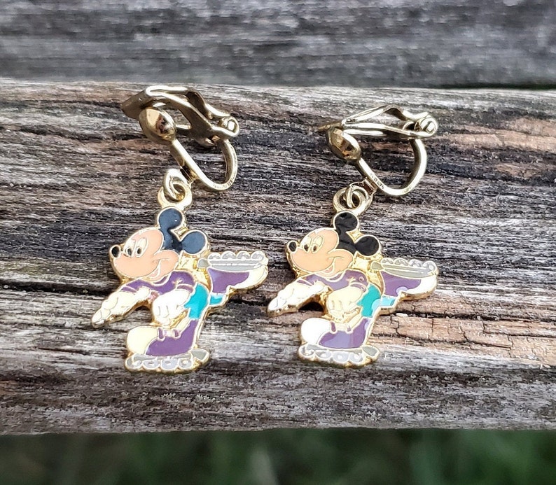 K&ouml;nnte beinhalten: Ein Paar goldfarbene Ohrclips mit einem Cartoon-Bild von Mickey Mouse beim Eislaufen. Die Ohrringe sind mit lila, t&uuml;rkis und schwarzem Email verziert.
