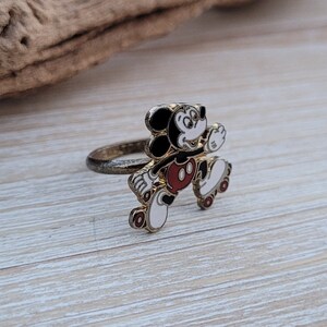 May include: Gold-tone ring featuring a cartoon Mickey Mouse on roller skates. Mickey is wearing red shorts and white gloves. The ring is a fun and whimsical accessory.