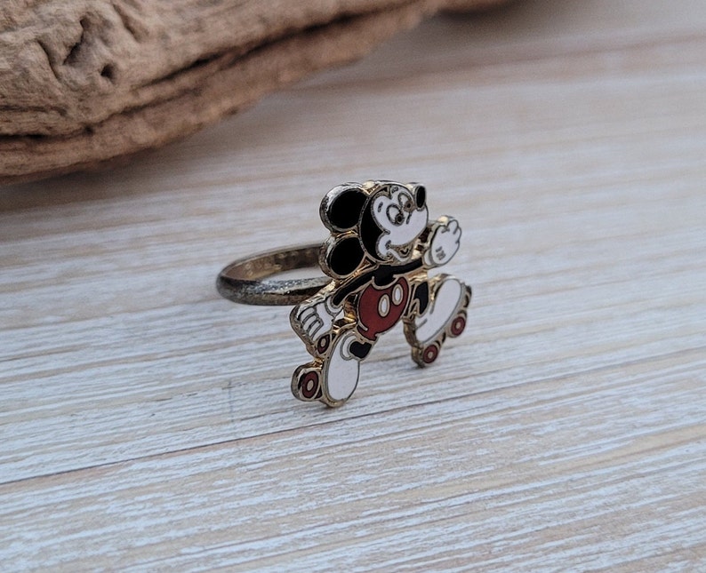 May include: Gold-tone ring featuring a cartoon Mickey Mouse on roller skates. Mickey is wearing red shorts and white gloves. The ring is a fun and whimsical accessory.