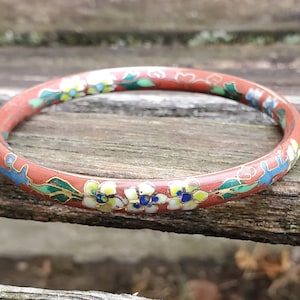 May include: A red enamel bangle bracelet with a floral design. The flowers are white with yellow centers and blue accents. The bracelet is decorated with green leaves and gold accents.