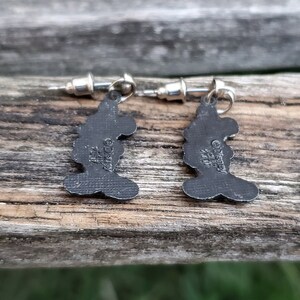 May include: A pair of silver-toned stud earrings featuring a silhouette of a character's head. The earrings are dark gray with a textured surface and have the text "&copy;Disney" and other markings.