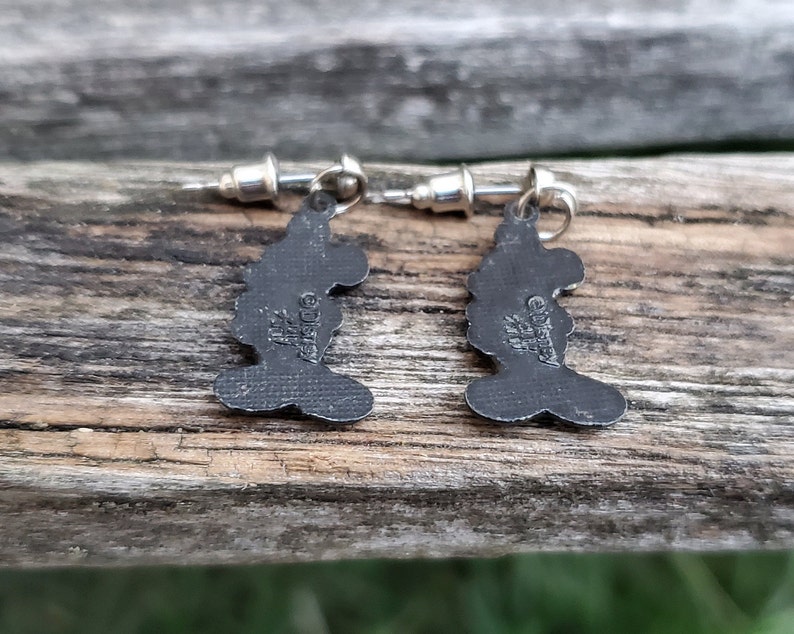 May include: A pair of silver-toned stud earrings featuring a silhouette of a character's head. The earrings are dark gray with a textured surface and have the text "&copy;Disney" and other markings.