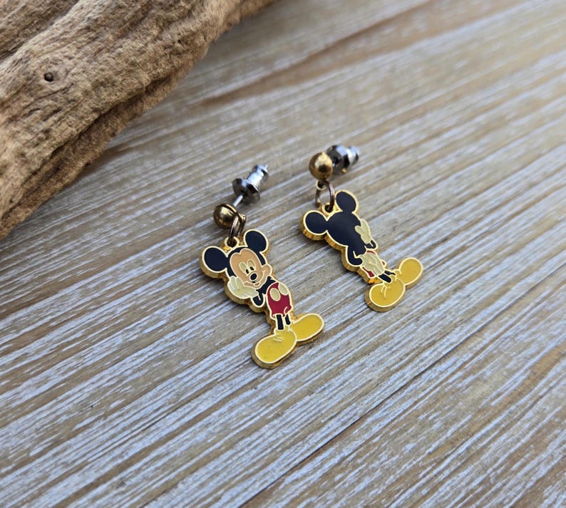 Puede incluir: Un par de pendientes de Mickey Mouse de color dorado. Los pendientes presentan una imagen de dibujos animados de Mickey Mouse con orejas negras, pantalones cortos rojos y zapatos amarillos. Los pendientes est&aacute;n unidos a postes de plata.