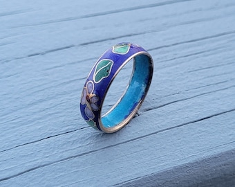 May include: A blue enamel ring with floral designs in green, purple, and white. The ring's interior is a bright turquoise color. The ring is set against a light blue wooden surface.