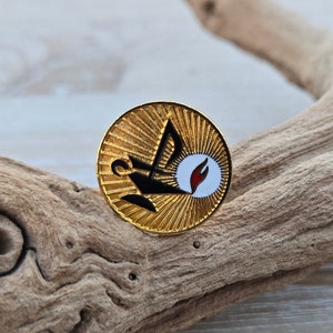 Könnte beinhalten: Goldfarbene runde Anstecknadel mit einem schwarz-weißen Emblem mit einer Lampe und einer Flamme. Die Anstecknadel hat ein Sonnenstrahlendesign um das Emblem. Die Anstecknadel befindet sich vor einem natürlichen Holzuntergrund.
