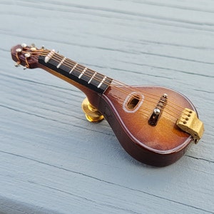 May include: A miniature wooden mandolin with a brown stain finish and gold hardware. The mandolin has a white inlay around the sound hole and a gold tuning peg.