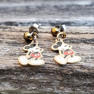 Puede incluir: Un par de pendientes de Mickey Mouse de color dorado. Cada pendiente tiene una peque&ntilde;a perla redonda dorada en la parte superior y un dije de Mickey Mouse con pantalones cortos rojos y zapatos negros.