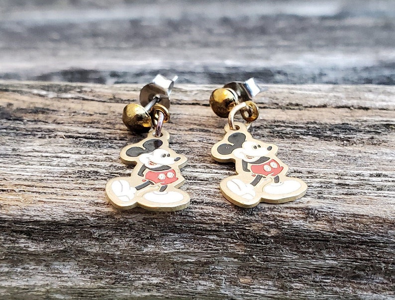 Puede incluir: Un par de pendientes de Mickey Mouse de color dorado. Cada pendiente tiene una peque&ntilde;a perla redonda dorada en la parte superior y un dije de Mickey Mouse con pantalones cortos rojos y zapatos negros.