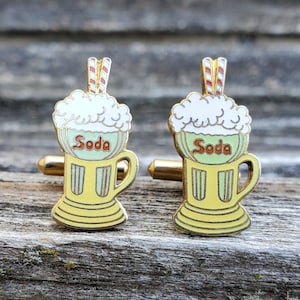 May include: A pair of gold cufflinks featuring a soda glass with a yellow base, white foam, and red and white straws. The glass says "Soda" in red letters.