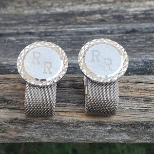 May include: A pair of silver cufflinks with a mesh band. The cufflinks have a circular design with a raised edge and the initials "R R" engraved on the front.