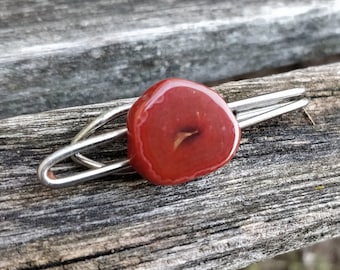 May include: A red gemstone tie clip with a silver metal clasp. The gemstone is oval shaped and has a smooth, polished surface.