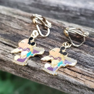 K&ouml;nnte beinhalten: Ein Paar goldfarbene Ohrclips mit einem Cartoon-Mickey-Mouse-Charakter, der lila Rollschuhe tr&auml;gt. Die Ohrringe sind mit buntem Email verziert.