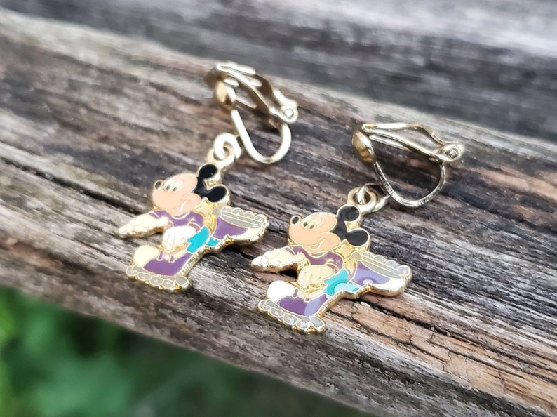 K&ouml;nnte beinhalten: Ein Paar goldfarbene Ohrclips mit einem Cartoon-Mickey-Mouse-Charakter, der lila Rollschuhe tr&auml;gt. Die Ohrringe sind mit buntem Email verziert.