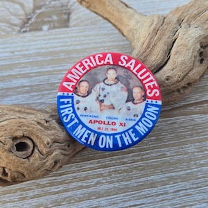 May include: A red, white, and blue button with a picture of the Apollo 11 astronauts. The text reads "America Salutes First Men On The Moon" and "Apollo XI July 20, 1969".