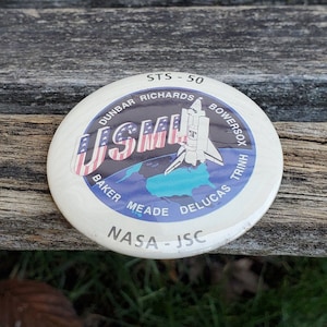May include: A white button with a blue and red US flag, a space shuttle, and the text "STS-50 NASA-JSC" and the names of the astronauts: Dunbar, Richards, Bowersox, Trinh, Baker, Meade, Delucas.