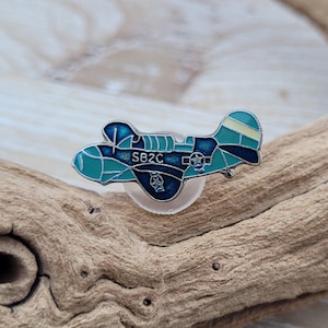 Pode incluir: Um broche de esmalte azul e verde de um avião vintage com o texto "SB2C" no lado. O broche está sobre uma superfície de madeira.