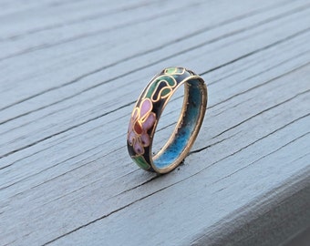 May include: A black ring with a floral design in pink, green, and blue enamel. The ring is made of metal and has a gold-colored band.