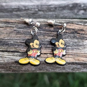 May include: A pair of dangle earrings featuring the iconic Mickey Mouse character. Each earring has a black, yellow, red, and white enamel design of Mickey Mouse. The earrings have silver-colored posts and are displayed on a weathered wooden surface.