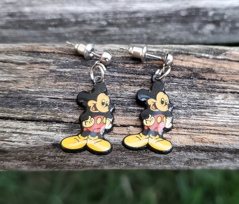 May include: A pair of dangle earrings featuring the iconic Mickey Mouse character. Each earring has a black, yellow, red, and white enamel design of Mickey Mouse. The earrings have silver-colored posts and are displayed on a weathered wooden surface.