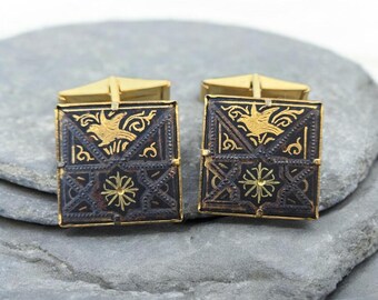 May include: A pair of square cufflinks with a gold-colored frame and backing. The face of each cufflink is dark brown with gold-colored decorative elements, including a bird and floral design. The cufflinks are displayed on a gray stone surface.