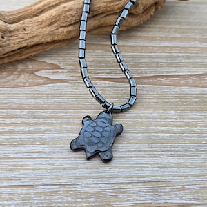 May include: A black hematite necklace with a black turtle pendant. The necklace is made of small, rectangular beads.