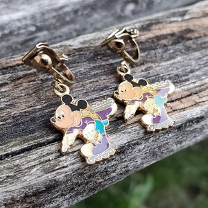 K&ouml;nnte beinhalten: Ein Paar goldfarbene Ohrclips mit Mickey Mouse und Minnie Mouse beim Rollschuhlaufen. Die Ohrringe sind mit buntem Email verziert und haben ein lila, gr&uuml;nes und gelbes Farbschema.